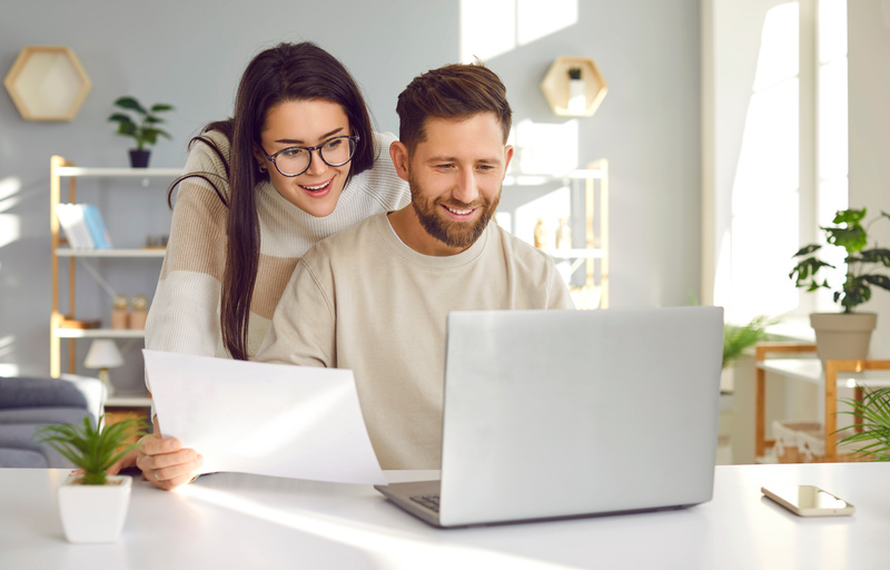 Cheerful young couple using laptop and analyzing their finances with paper document. Smiling husband and wife using online banking service at home or receiving positive decision from bank by email